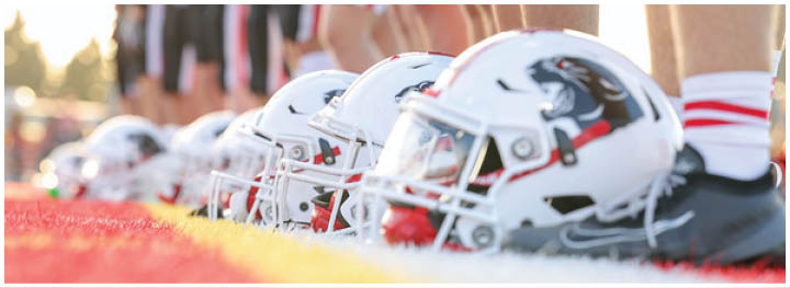 A row of football helmets is lined up on the ground in front of people standing in athletic gear on a field during sunset. Description generated by AI