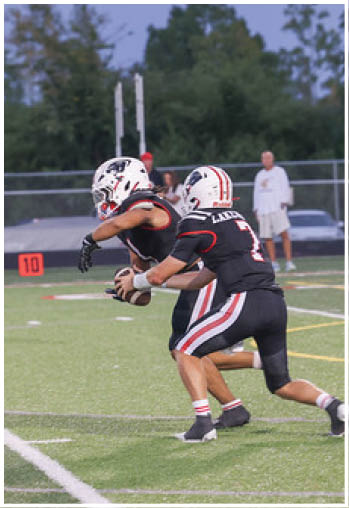 Two people in football uniforms and helmets are on a field with one handing off a football to the other during a game. Description generated by AI