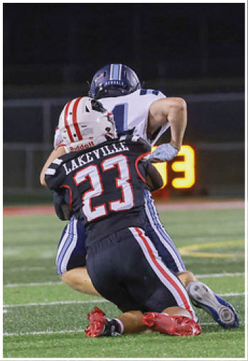 A person wearing a black football uniform with the name Lakeville and number 23 is tackling another person in a white and blue football uniform on a field at night. Description generated by AI