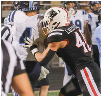 Two people in football uniforms are engaged in a play on the field with one person in black and red attempting to block or tackle the other person in white and blue. Description generated by AI