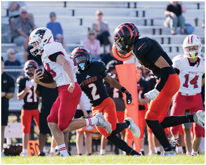 A person in a white and red football uniform is running with the ball while two people in black and orange uniforms chase them on a grassy field with spectators in the background. Description generated by AI