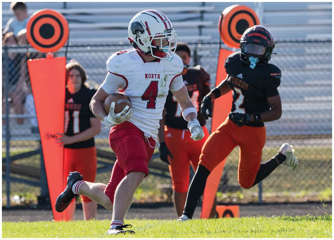 A person in a white and red football uniform is running with the ball while another person in a black and orange uniform chases behind on a grassy field with bleachers and spectators in the background. Description generated by AI
