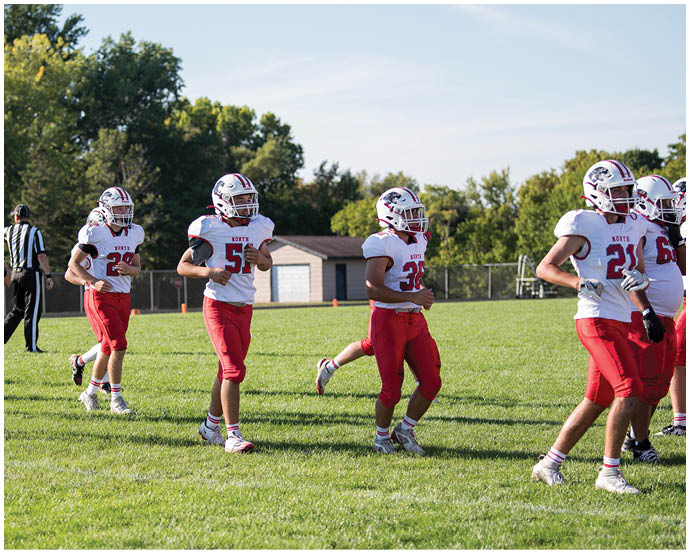Several people wearing white helmets and red sports uniforms are walking on a grassy field with trees and a small building in the background. Description generated by AI