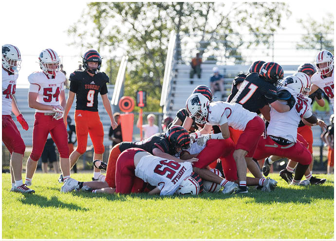 A group of people wearing football uniforms are engaged in a play on a grassy field with some standing and others piled on the ground. Description generated by AI