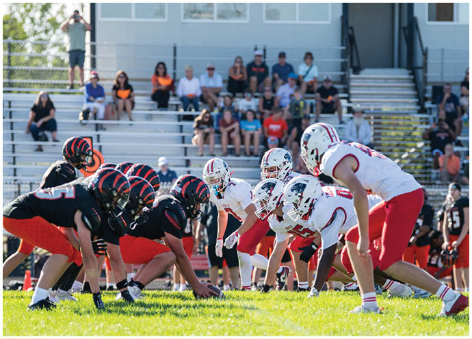 Two teams of adults in football uniforms are lined up facing each other on a grassy field with spectators seated on bleachers in the background. Description generated by AI