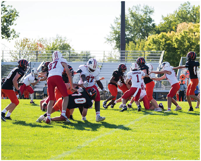 A group of people wearing football uniforms are actively engaged in a play on a grassy field with bleachers and trees in the background. Description generated by AI