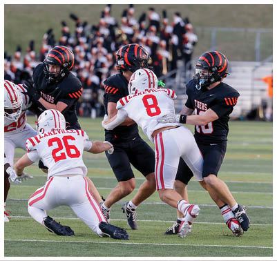 Several people in football uniforms are engaged in a play on a grassy field with a group of band members in the background. Description generated by AI