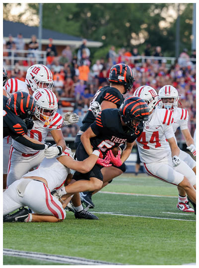 A group of football players in black and white uniforms are engaged in a tackle on a grassy field during a game with a crowd in the background. Description generated by AI