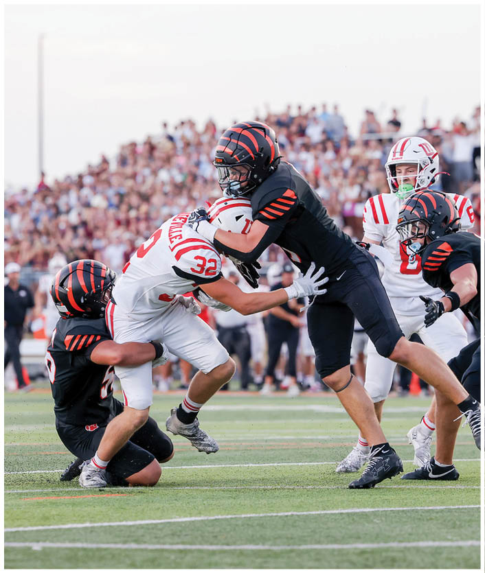Four football players in black uniforms are tackling a player in a white uniform on a field with a crowd in the background. Description generated by AI