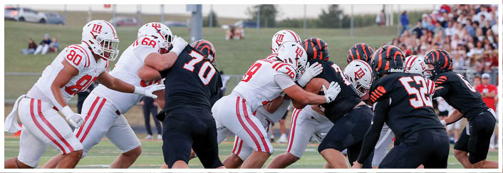Two opposing American football teams in white and black uniforms are engaged in a play on a field with a crowd of spectators in the background. Description generated by AI