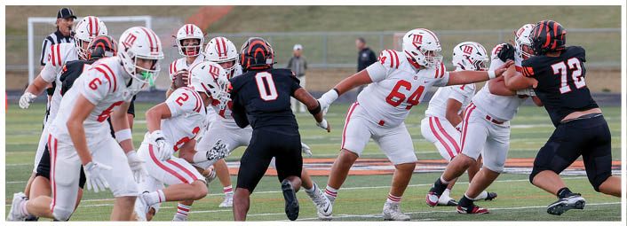 Two opposing American football teams in white and black uniforms are engaged in a play on a grassy field with players blocking and preparing to advance. Description generated by AI