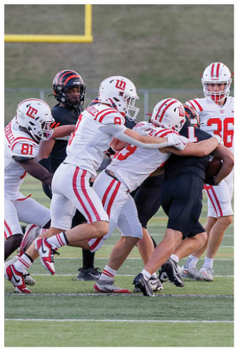Several people in white and red football uniforms are tackling a person in a black football uniform on a green field with a grassy hill and parked cars in the background. Description generated by AI
