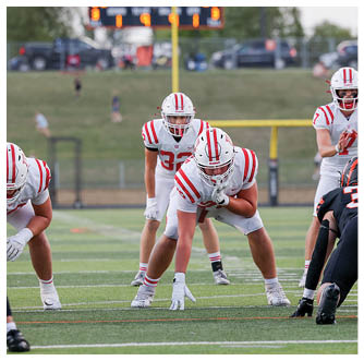 Several people in football uniforms are positioned on a field preparing for a play, with one person holding the ball and another facing them in a defensive stance. Description generated by AI