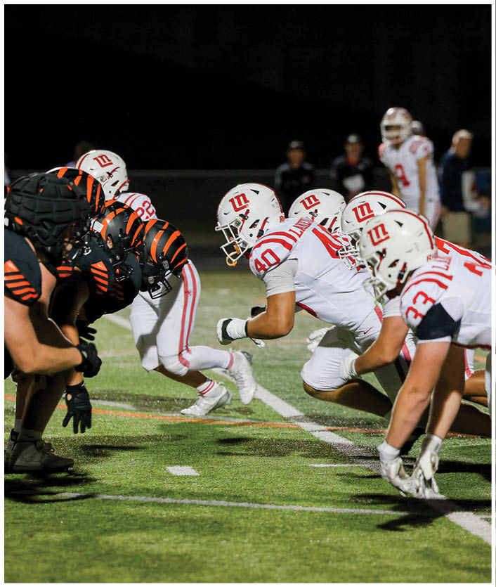 Two opposing American football teams in black and orange and white and red uniforms are lined up facing each other on a field under stadium lights. Description generated by AI