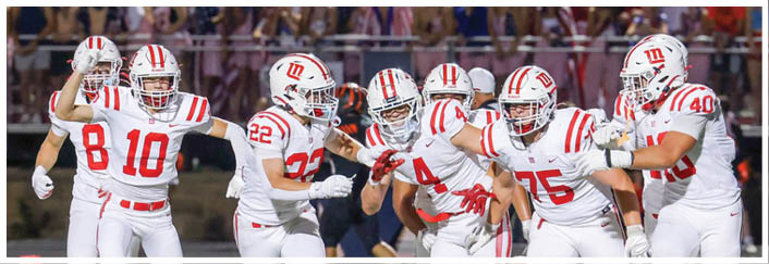 A group of people in white sports uniforms with red stripes are celebrating on a football field while a person in a black uniform walks away in the background. Description generated by AI