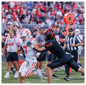 A person in a white football uniform is running with the ball while another person in a black football uniform reaches out to tackle them on a field with spectators in the background. Description generated by AI