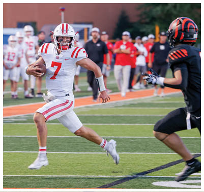 A person wearing a white football uniform with red stripes and the number seven is running on a football field while holding a football, with another player in a black and orange uniform approaching. Description generated by AI