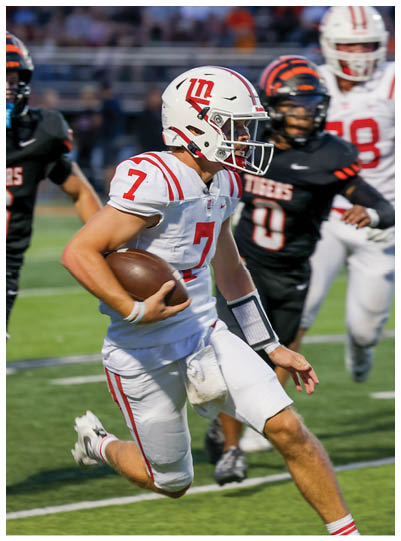 A person wearing a white football uniform with red stripes and the number seven is running with a football while being pursued by players in black and white uniforms on a grassy field. Description generated by AI