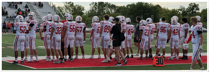 A group of people wearing white and red football uniforms stand on the sidelines of a football field with spectators seated in the bleachers in the background. Description generated by AI