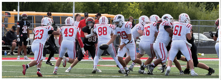 A group of people in white and red football uniforms are engaged in a play on a football field with coaches and other team members in the background. Description generated by AI