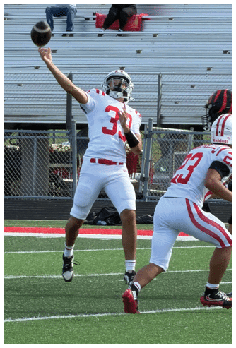 Two people in football uniforms are on a field with one preparing to throw a football while the other is in a ready stance, and two people are seated in the bleachers behind them. Description generated by AI