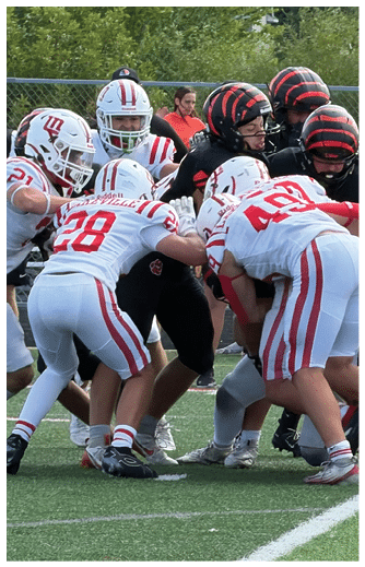 A group of people wearing football uniforms are engaged in a play on a grassy field with a fence and trees in the background. Description generated by AI