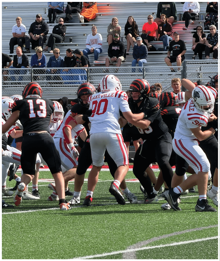 Adults in football uniforms are engaged in a game on a green field with spectators seated on bleachers in the background. Description generated by AI