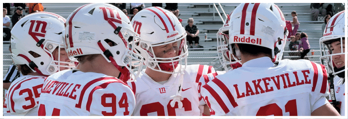 People wearing white and red football helmets and jerseys with “Lakeville" on the back are gathered closely, likely during a game or practice. Description generated by AI