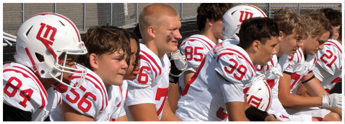 Several people wearing white and red football uniforms and helmets are sitting on a bench, appearing focused and attentive. Description generated by AI
