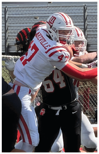 Players in white and black football uniforms are engaged in a play on a green field with bleachers and spectators in the background. Description generated by AI