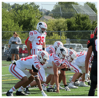 A group of people wearing white and red football uniforms are lined up on a field preparing for a play with a person in casual clothing standing nearby holding a tablet. Description generated by AI