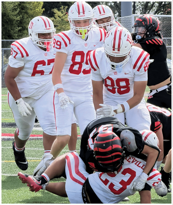 Several people in white and red football uniforms are standing on a field while two others in black and orange uniforms are on the ground in a tackle. Description generated by AI