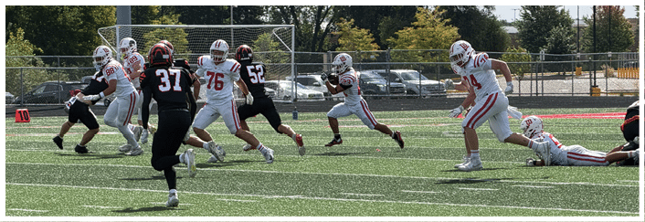 People in sports uniforms are running and preparing to play on a grassy field with a chain-link fence and parked cars in the background. Description generated by AI