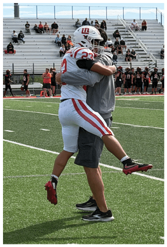 A person in a football uniform jumps into the arms of another person wearing casual clothing on a football field with spectators in the background. Description generated by AI