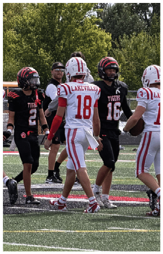 Adults wearing football uniforms from two different teams are standing and interacting on a football field during a game. Description generated by AI