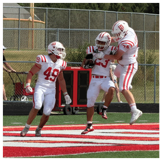 Three people in football uniforms are on a field with one holding a football while a person in casual clothing pushes a stroller behind a fence in the background. Description generated by AI