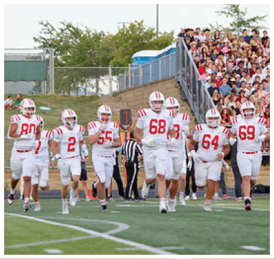 A group of football players in white uniforms with red stripes are running onto a field with a large crowd of people watching from the stands in the background. Description generated by AI