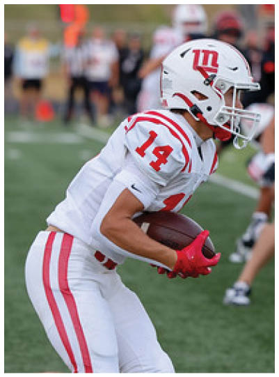 A person wearing a white football uniform with red stripes and the number 14 is running on a football field while holding a football. Description generated by AI
