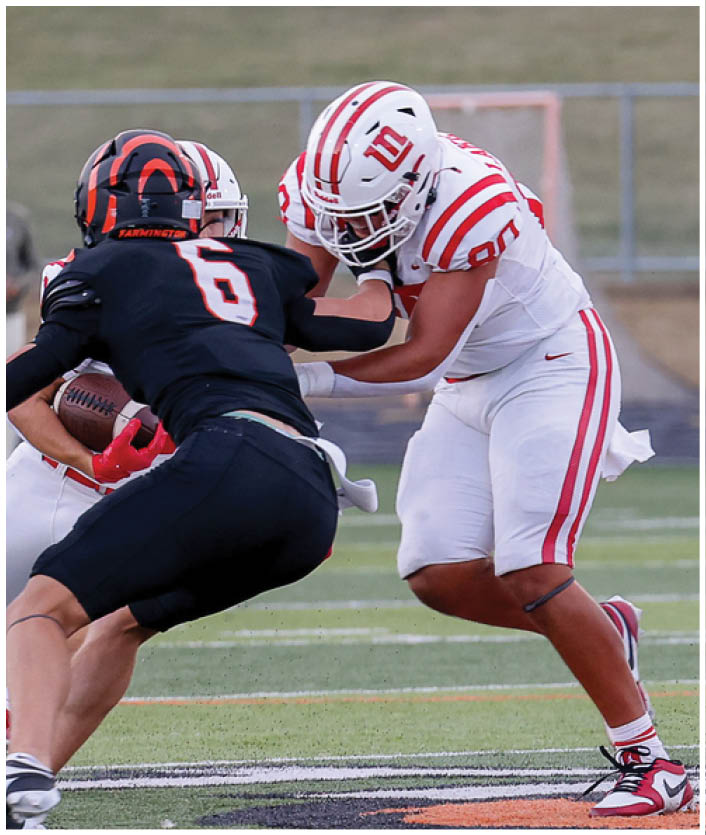 Two people in football uniforms are engaged in a play on a field, with one person holding the ball and the other attempting to block or tackle. Description generated by AI