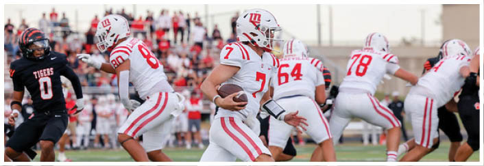 People in white and black sports uniforms are playing football on a field with one person in white running with the ball while others prepare to block or tackle. Description generated by AI