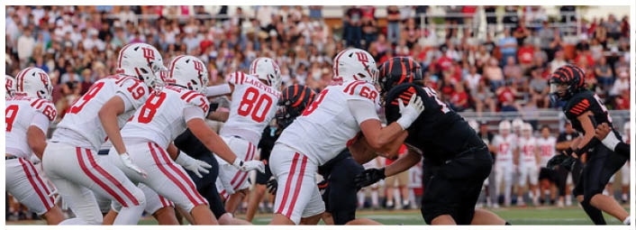 People in football uniforms from two opposing teams are engaged in a play on a crowded field with spectators in the background. Description generated by AI