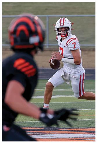 A person in a white football uniform is preparing to throw a football while another person in a black and orange uniform is in the foreground on a football field. Description generated by AI