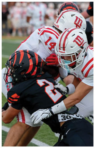 A person in a white and red football uniform is tackling a person in a black football uniform on a green field with other players and spectators in the background. Description generated by AI