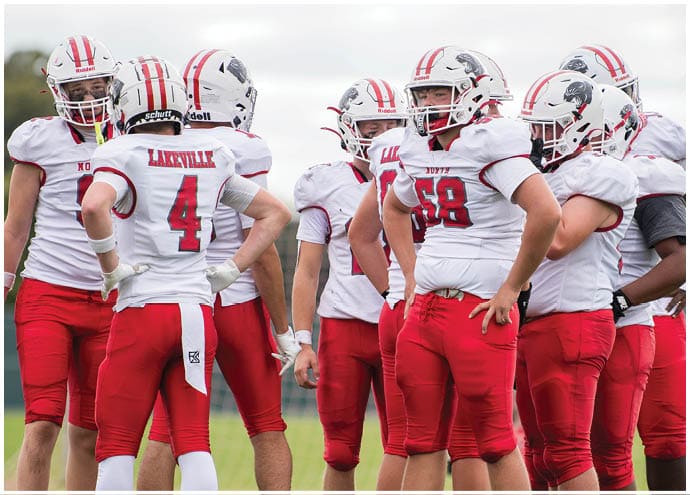 A group of people wearing white helmets, white jerseys, and red pants are standing together on a grassy field, likely during a football game. Description generated by AI
