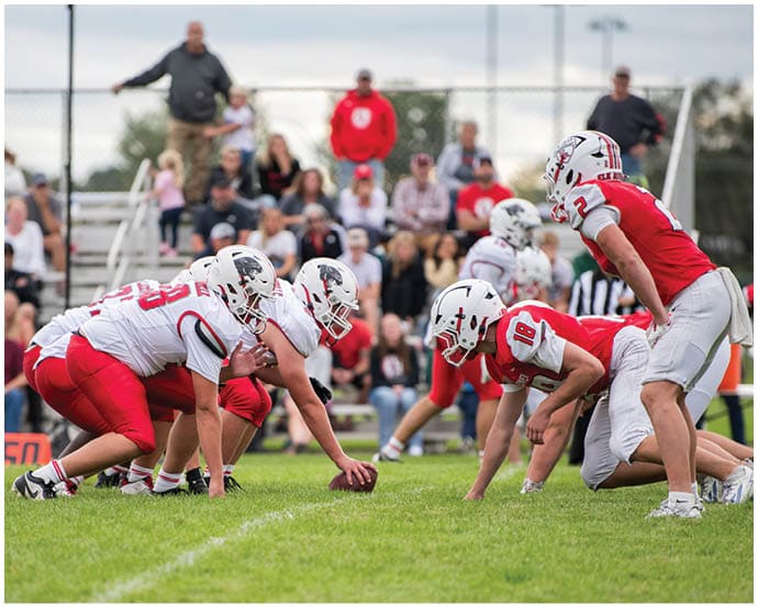 Two opposing American football teams in red and white uniforms are lined up facing each other on a grassy field preparing for a play. Description generated by AI
