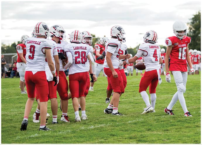 A group of people wearing white helmets and red and white football uniforms are gathered on a grassy field, with one person holding a football. Description generated by AI