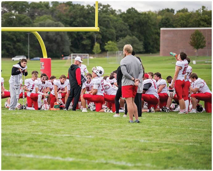A group of people in red and white football uniforms are gathered on a grassy field near a goalpost, with some standing and others sitting or kneeling while one person drinks from a bottle. Description generated by AI