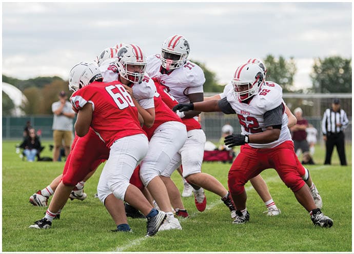 Several people wearing football helmets and uniforms are engaged in a play on a grassy field with a referee and spectators in the background. Description generated by AI