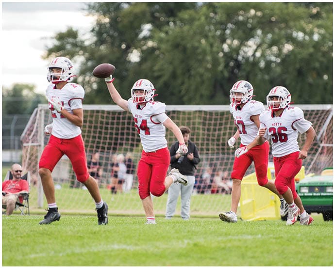 Four people in red and white football uniforms are running on a grassy field with one person holding a football in the air near a goal net. Description generated by AI