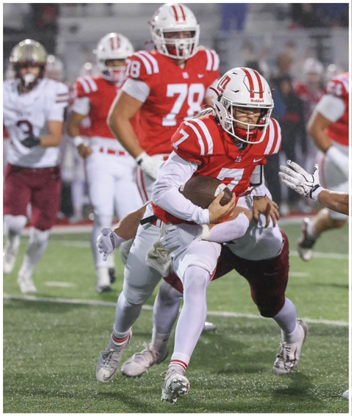 A person in a red and white football uniform is running with the ball while being pursued by a person in a white and maroon uniform on a football field with other players and spectators in the background. Description generated by AI